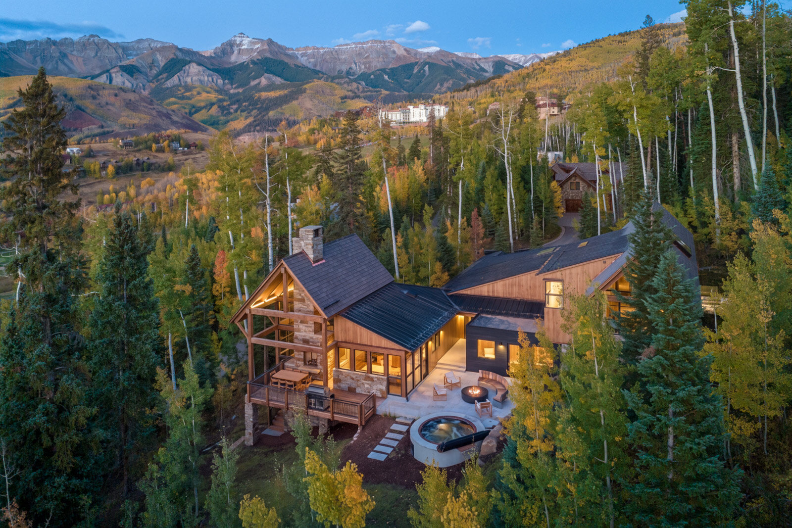 Aerial view of a timber mountain home among aspens and pines at golden hour, with the San Juan peaks beyond. Galloping Goose Chalet, Mountain Village, CO